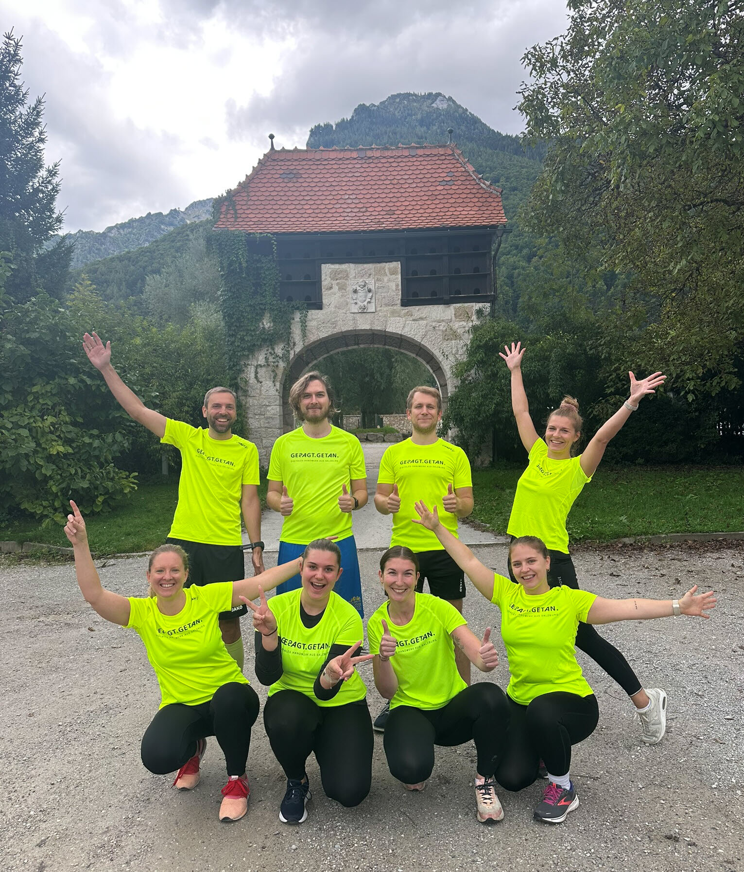 Acht Personen in neon-gelben T-Shirts posieren fröhlich vor einem historischen Tor, mit Bergen im Hintergrund. Teamfoto bei Outdoor-Event.