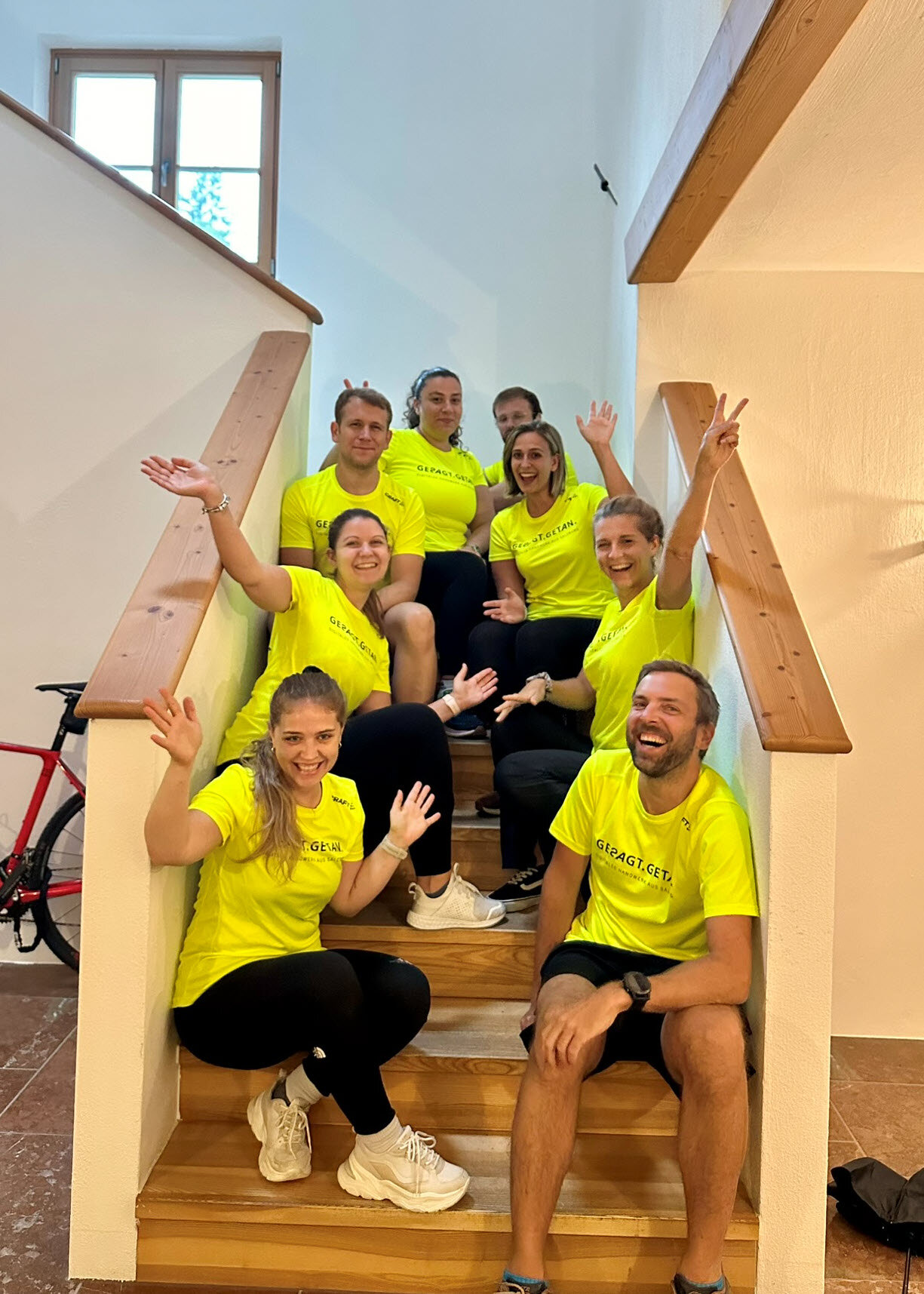 Gruppe in neon-gelben Teamshirts sitzt lachend und winkend auf einer Stiege im Haus und schaut in die Kamera.